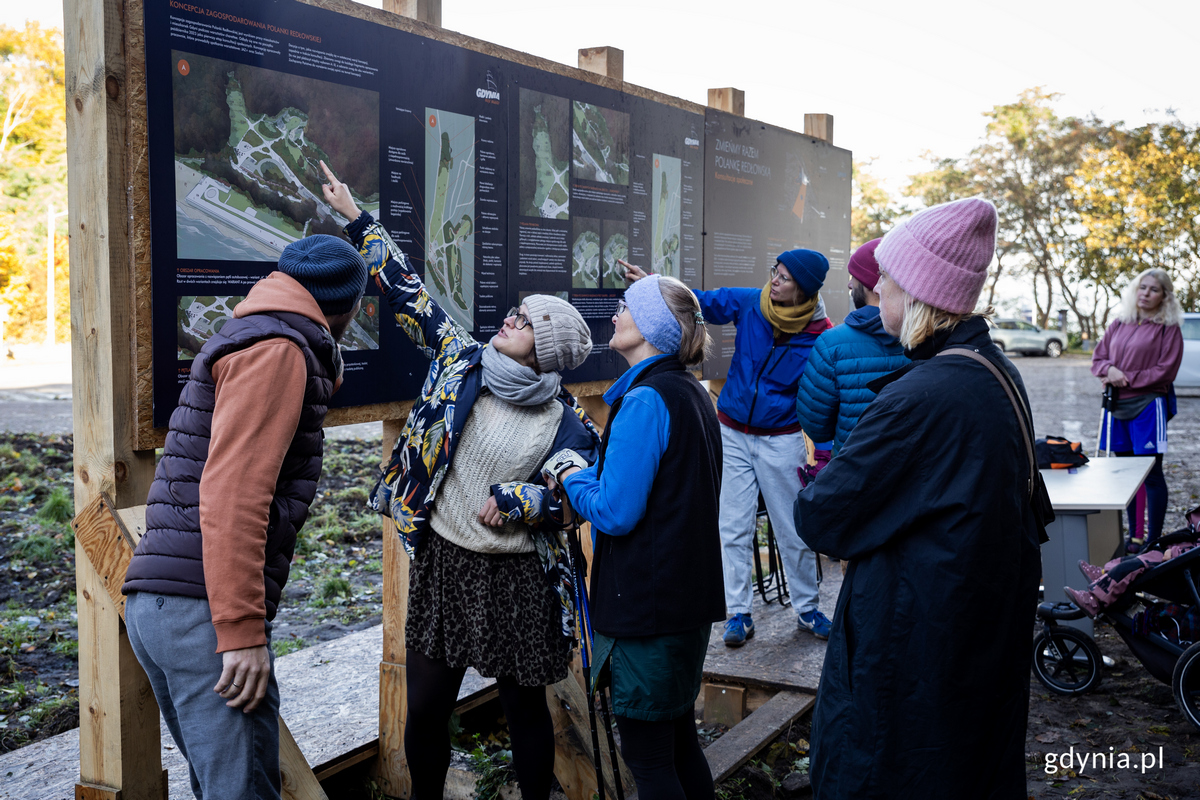 kilka os&oacute;b rozmawia przed tablica informacyjna na polanie.