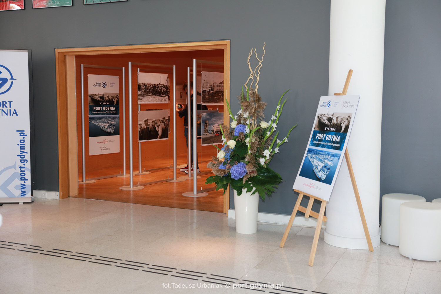 Plakat informujący o wystawie Port Gdynia w Muzeum Miasta. Na zdj. fragment muzealnej sali z zawieszonymi zdjęciami, fot. Tadeusz Urbaniak 