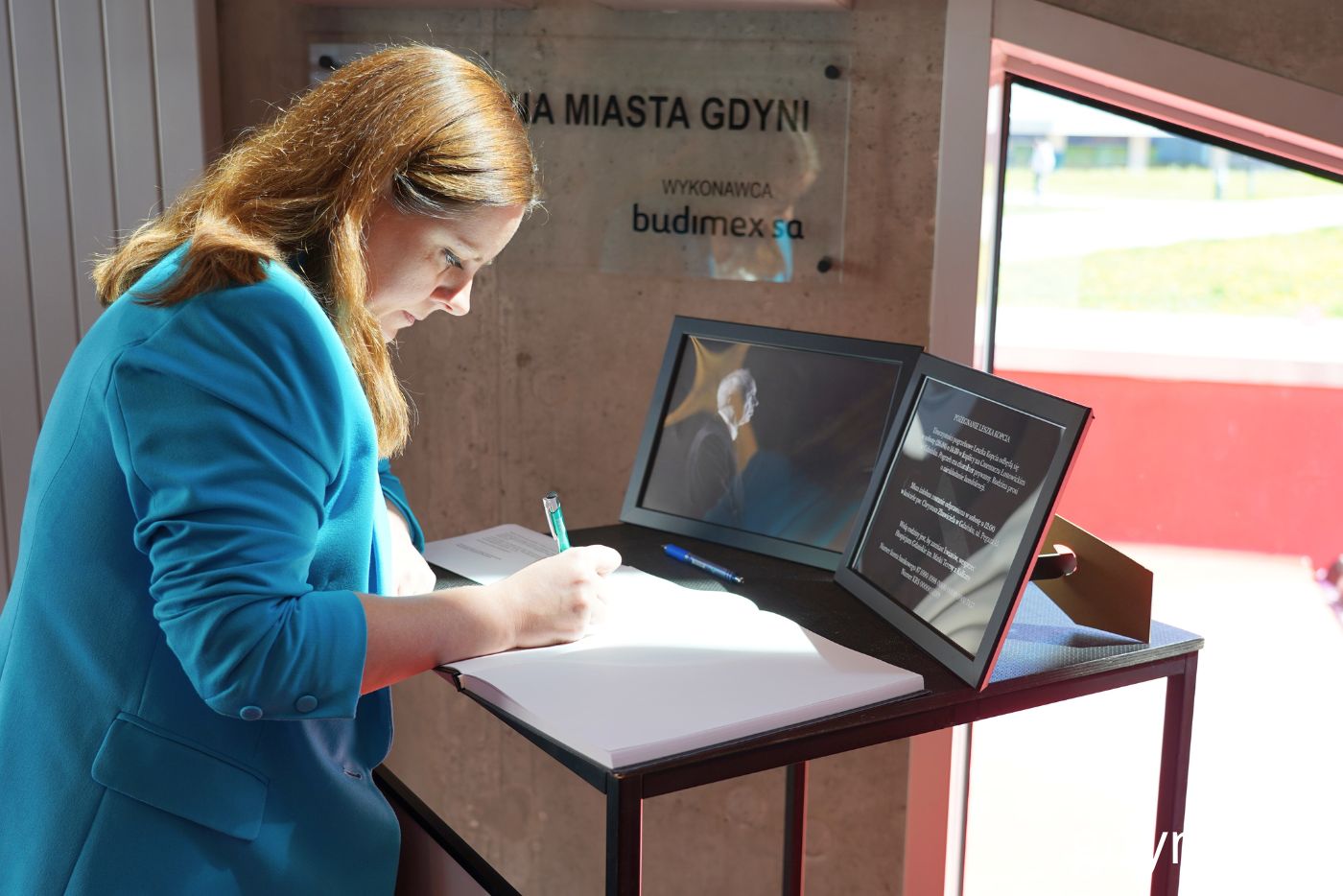Hol Gdyńskiego Centrum Filmowego, czarny stolik ze zdjęciem Leszka Kopcia i księgą kondolencyjną, stoi prezydent Aleksandra Kosiorek, wpisuje się do księgi