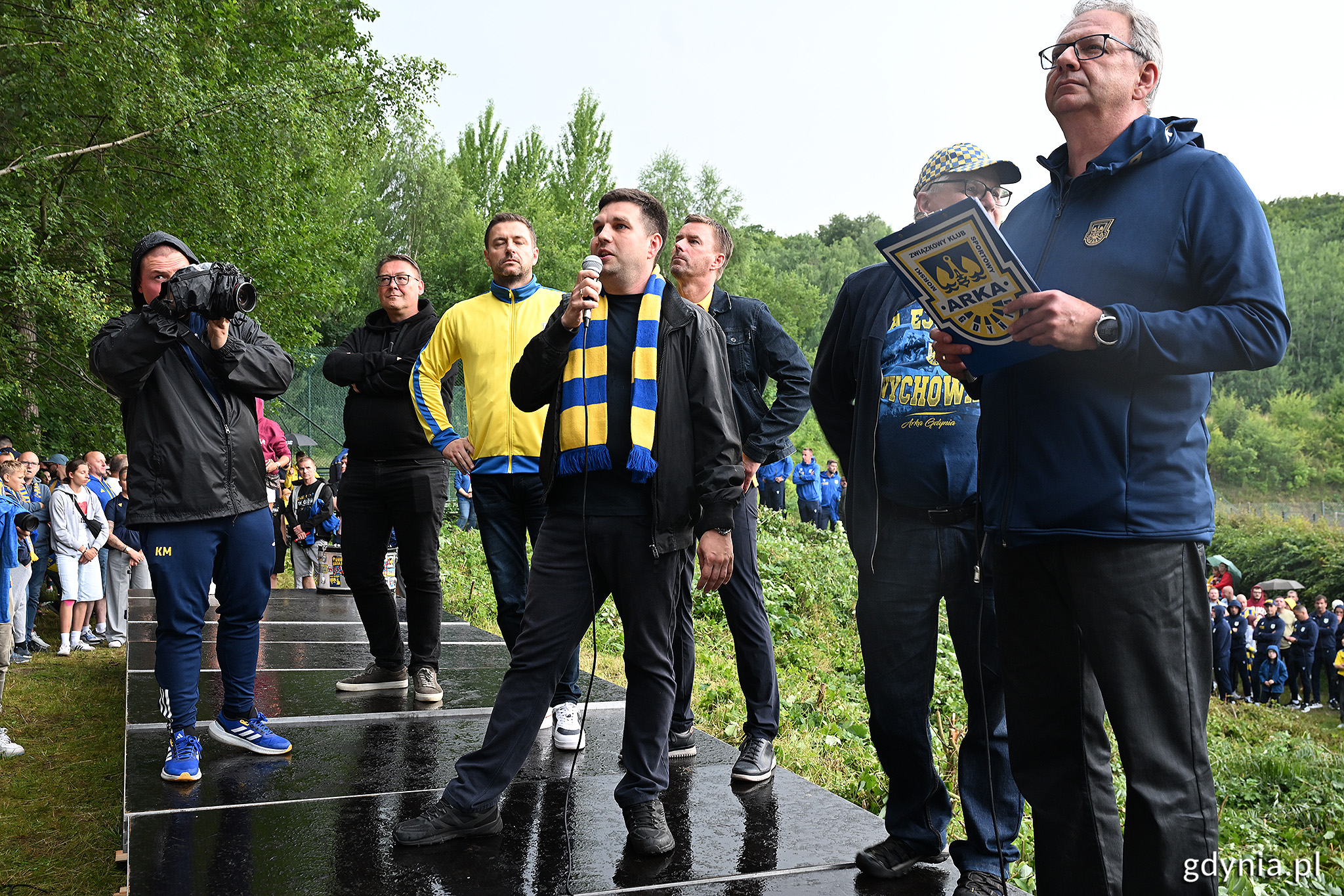 Platforma przy G&oacute;rce na ul. Ejsmonda, w tle korty tenisowe i las, na pierwszym planie grupa os&oacute;b, na środku Maciej Zielonka, kt&oacute;ry przemawia przez mikrofon