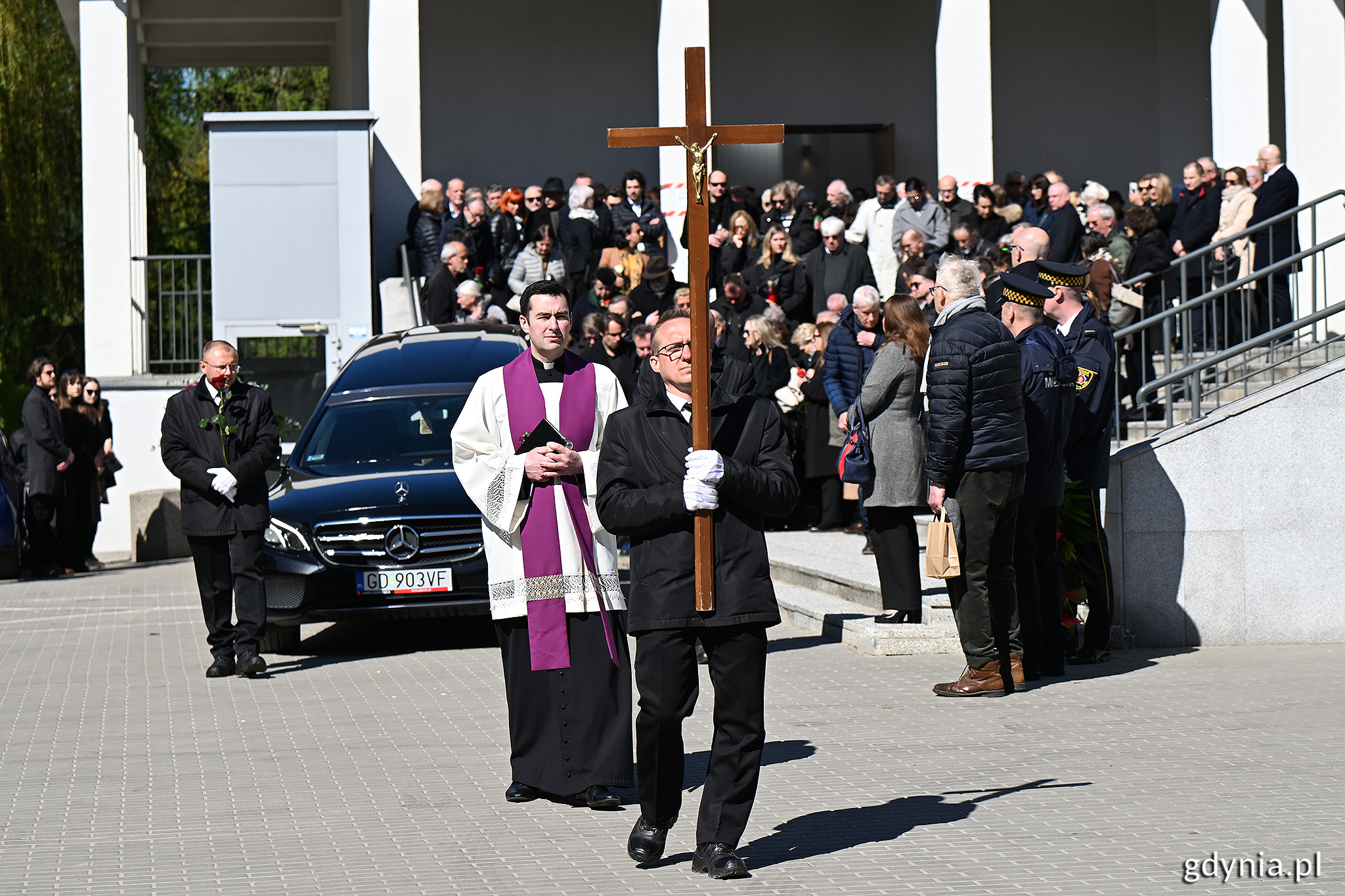 Kaplica na cmentarzu, tłum ludzi, samoch&oacute;d - karawan, na czele pochodu niesiony krzyż, za nim ksiądz