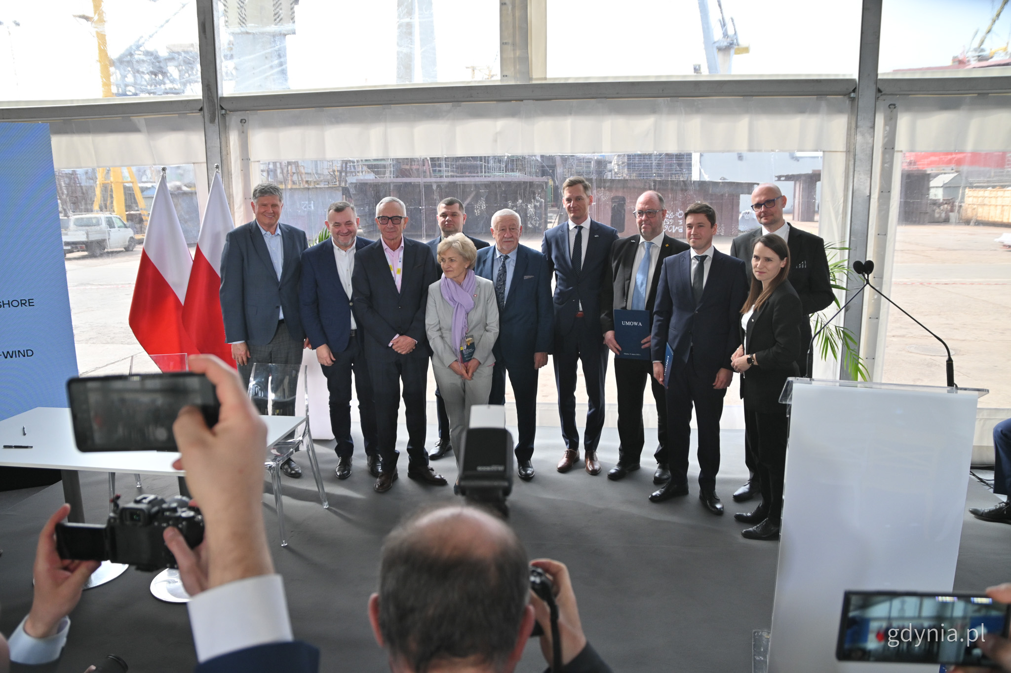 Sala konferencyjna, namiot, na scenie stoi grupa os&oacute;b - mężczyzn i kobiet - ubranych formalnie, pozują do zdjęcia, przed nimi fotoreporterzy