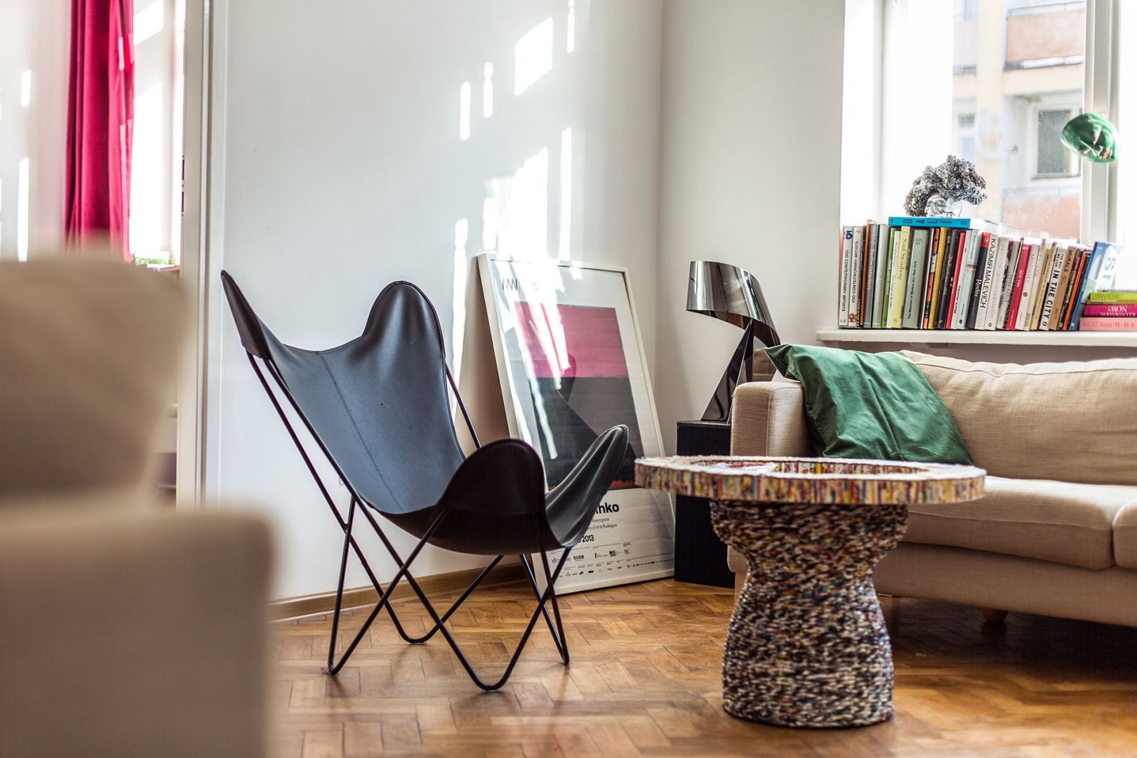 Wnętrze mieszkania Diany, a w nim czarne krzesło, ozdobny stolik, beżowa sofa z zieloną poduszką oraz książki poukładane na oknie. Na podłodze, oparta o ścianę, stoi grafika oprawiona w ramkę // fot. Dominik Jagodziński