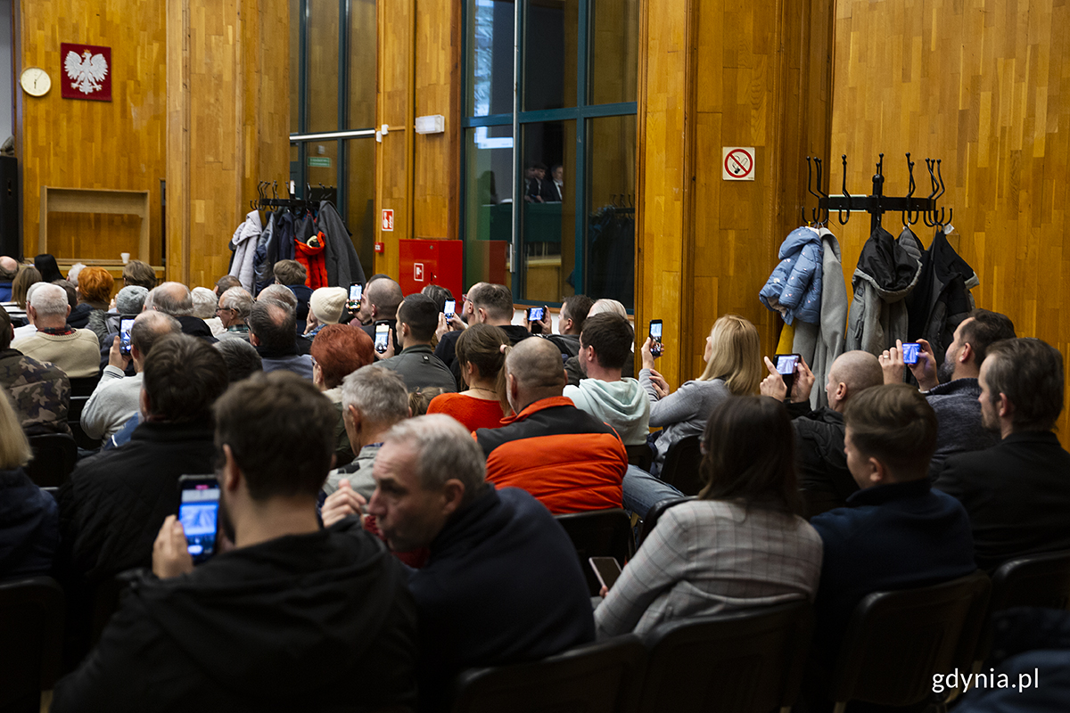 Ludzie widziani od tyłu siedzą na dużej sali i nagrywają prezentację telefonami.