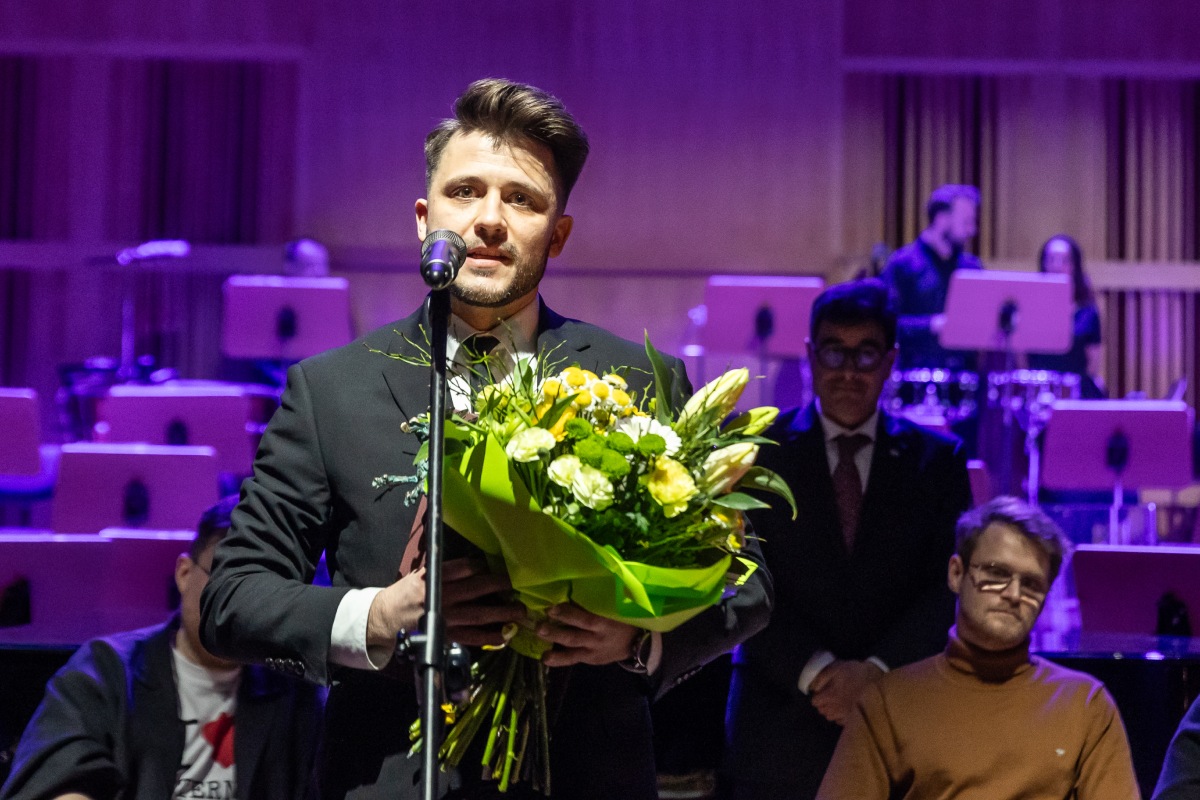 Saksofonista i kompozytor Szymon Zawodny podczas gali Pomorskiej Nagrody Artystycznej. Stoi przy mikrofonie, w rękach trzyma bukiet kwiat&oacute;w (fot. Piotr Żagiell)