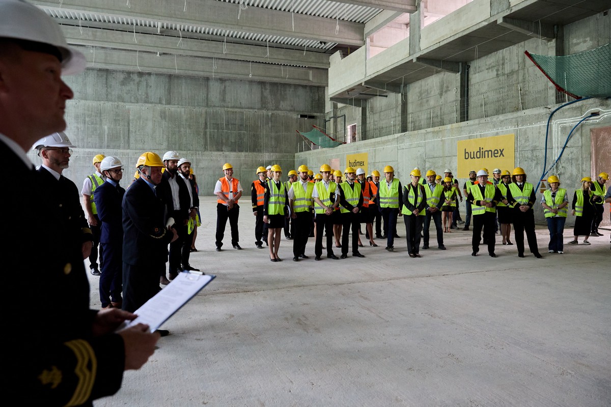 Uczestnicy uroczystości podniesienia wiechy na powstającym budynku Centrum Sportu i Rekreacji Uniwersytetu Morskiego w Gdyni zebrani wewnątrz hali. Mają na sobie odblaskowe kamizelki oraz kaski. Wśr&oacute;d nich Joanna Zielińska - przewodnicząca Rady Miasta Gdyni i przewodnicząca rady uczelni (druga od prawej w pierwszym rzędzie) // fot. Jacek Koślicki / Koslicki.pl