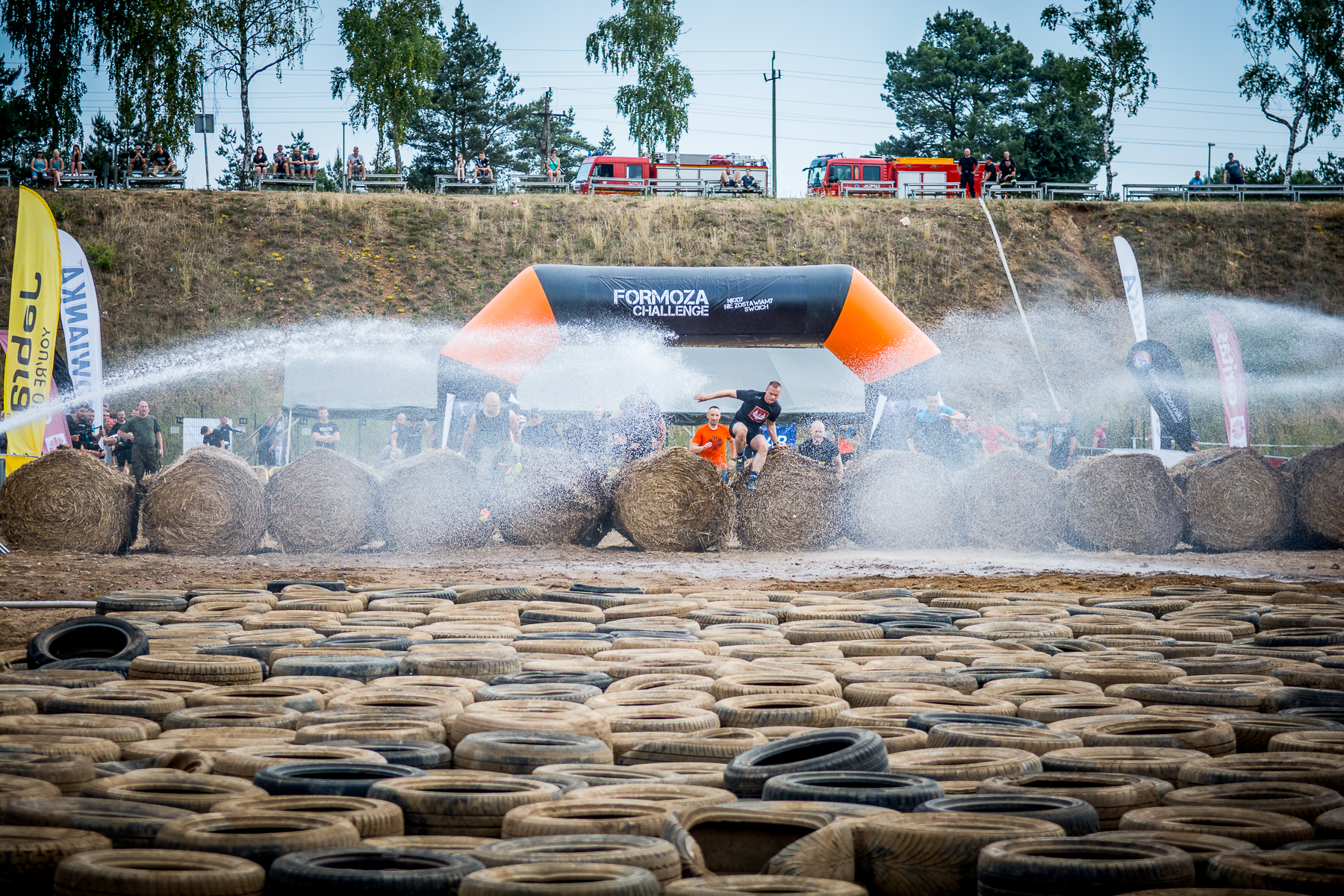 Tor przeszk&oacute;d na zawodach Formoza Challenge. Jeden z zawodnik&oacute;w przeskakuje przez ułożone w rzędzie bele siana, przed nim przeszkody w postaci gumowych opon. Po bokach, z obu stron, pryska woda // fot. materiały prasowe