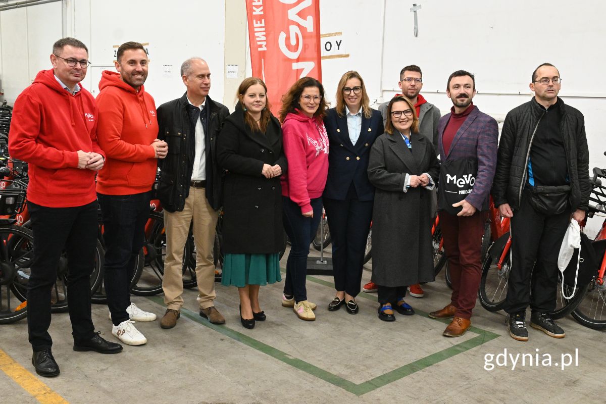 Grupa ludzi stojących w hali przed mikrofonami i mających za sobą rzędy ustawionych rower&oacute;w