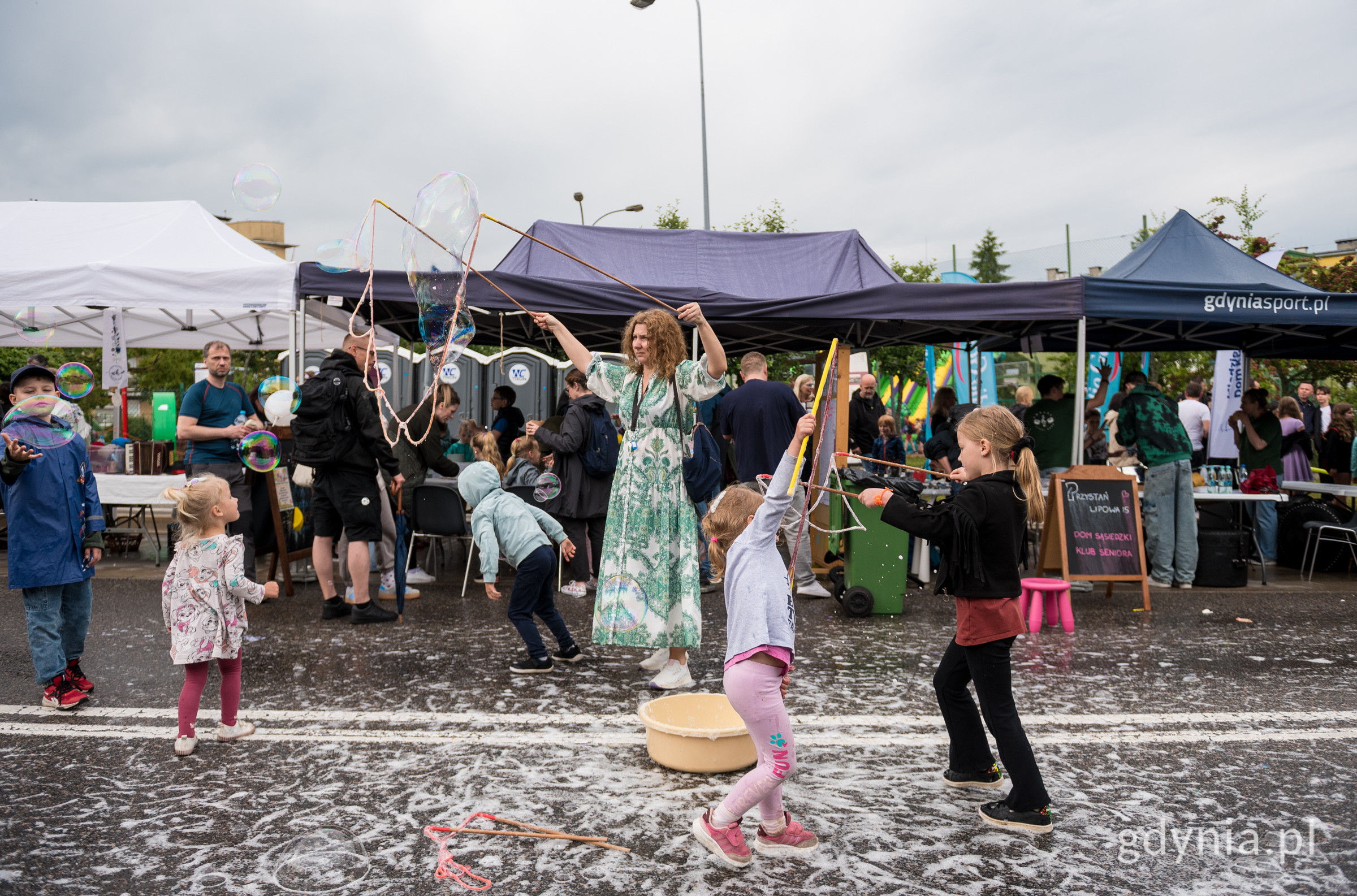 Chodnik, namioty w tle, festyn, zachmurzone niebo. Widoczny tłum ludzi, na pierwszym planie kobieta tworzy bańki mydlane, obok kilkoro dzieci