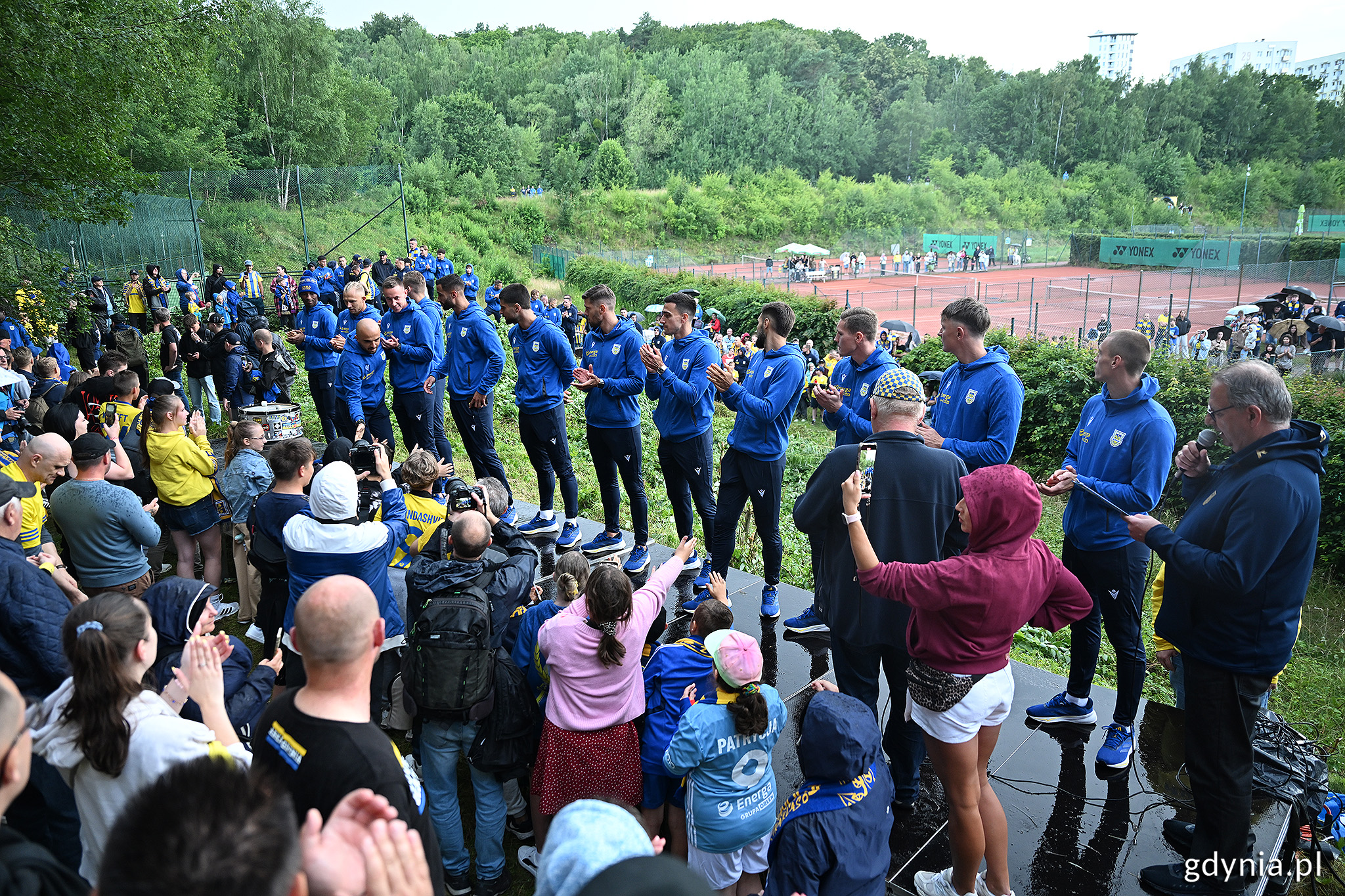 G&oacute;rka przy ul. Ejsmonda, w tle korty tenisowe i las, na pierwszym planie platforma, na kt&oacute;rej stoją piłkarze w niebieskich dresach