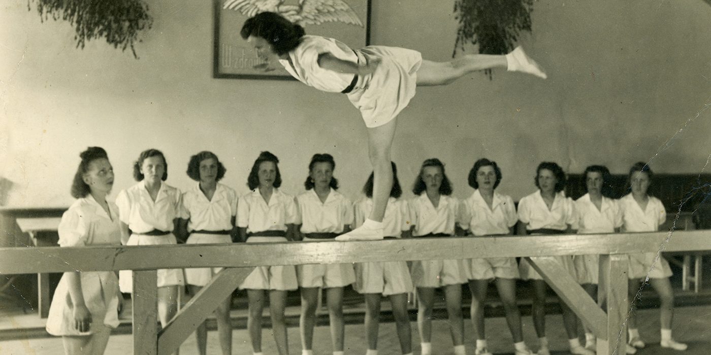Zdjęcie archiwalne wykonane w sepii. Grupa kobiet podczas ćwiczeń gimnastycznych. Jedna z nich wykonuje ćwiczenia na drewnianej platformie. 