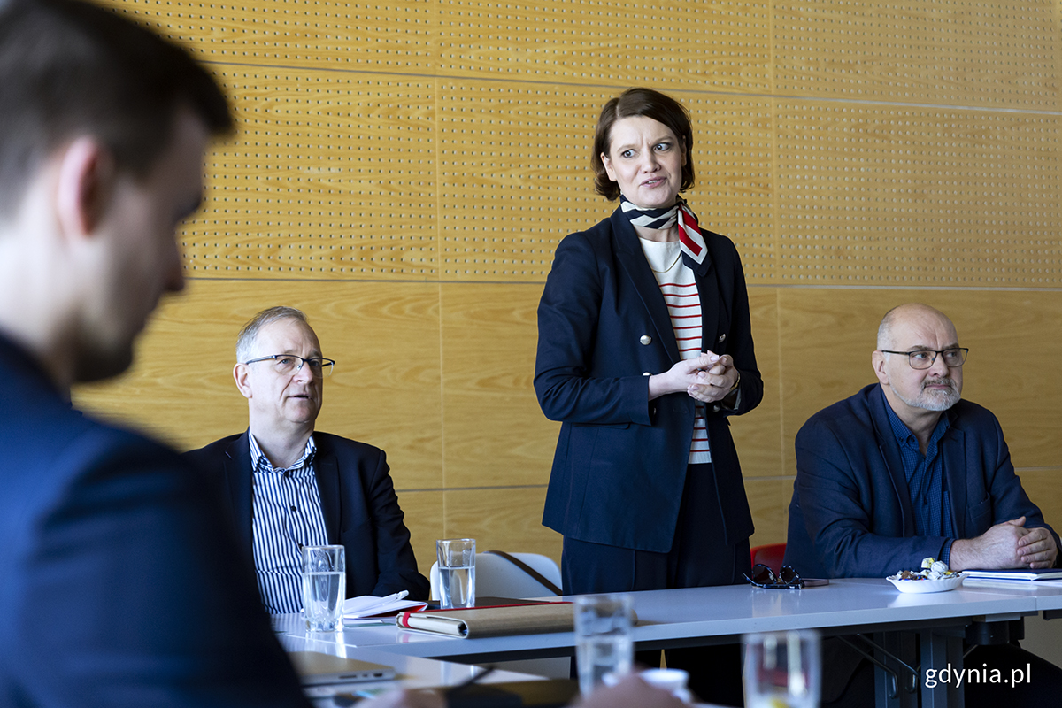 Kobieta stoi w sali seminaryjnej i m&oacute;wi, obok siedzi dw&oacute;ch mężczyzn.