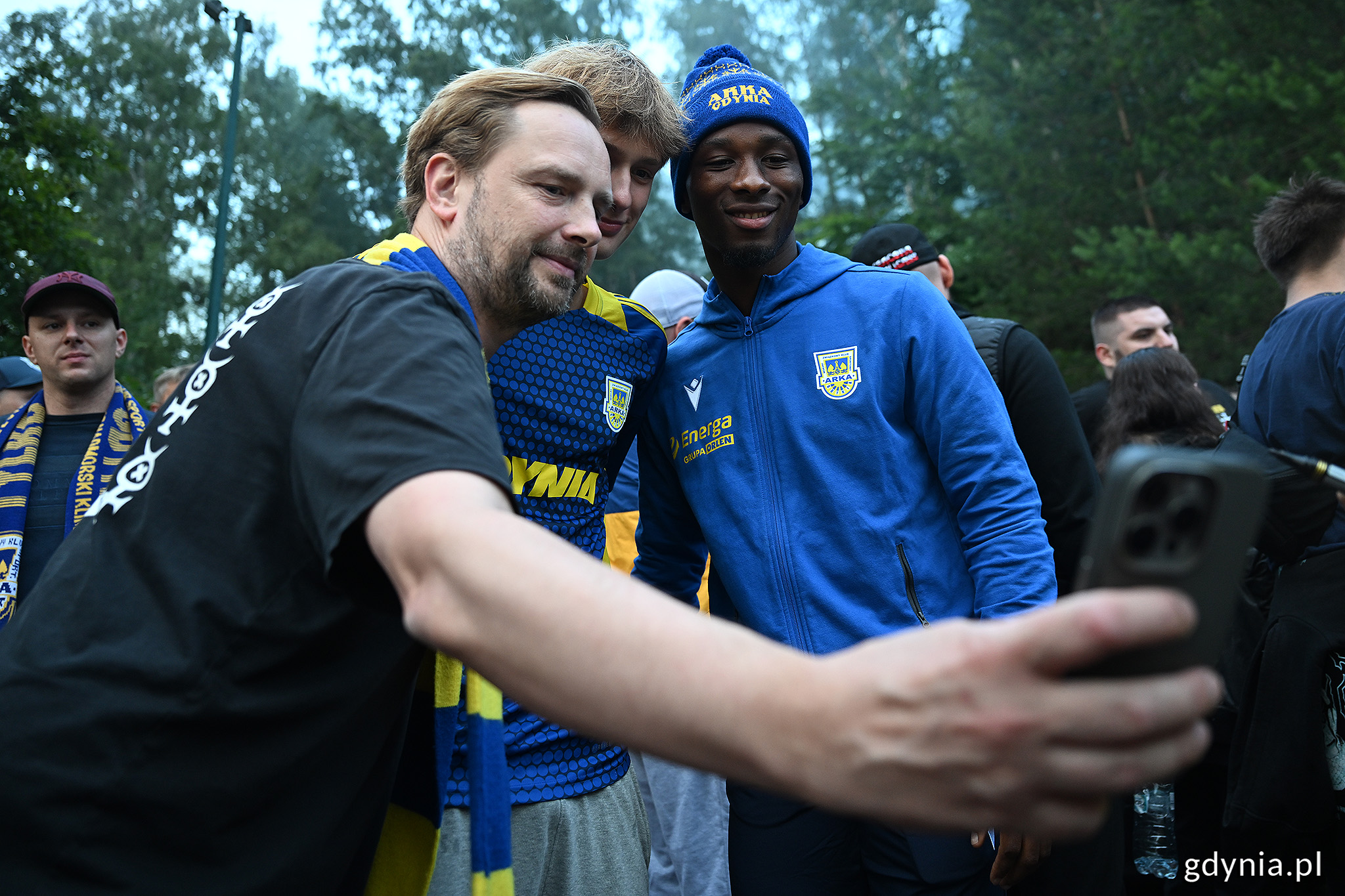 Las, G&oacute;rka przy Ejsmonda, tłum ludzi, mężczyzna i dziecko robią telefonem zdjęcie z piłkarzem Arki, Aurelienem Nguiambą