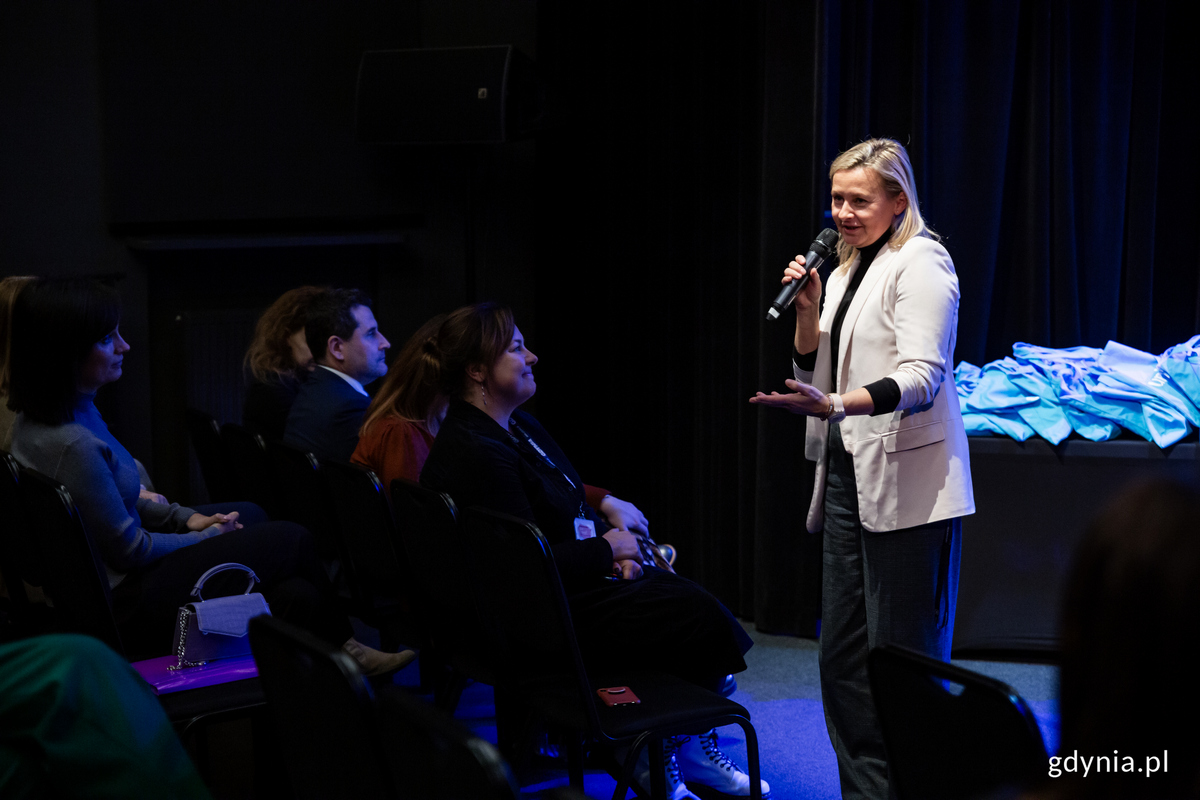 Kobieta stoi i m&oacute;wi do siedzących przed nia ludzi.