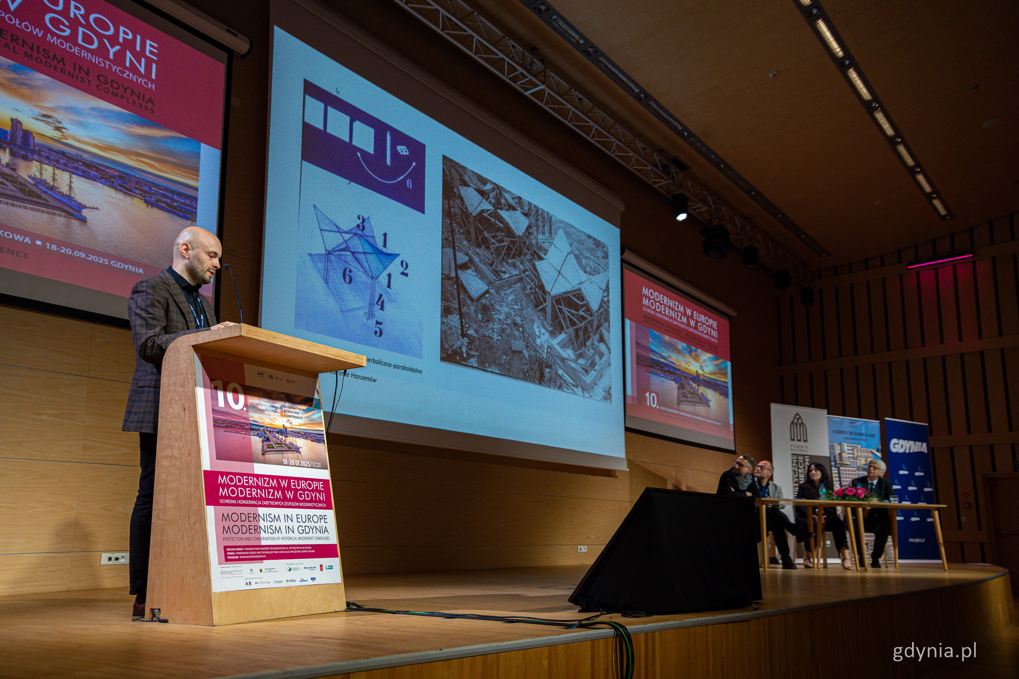 Sala konferencyjna w PPNT, scena, na ekranie zdjęcia architektury, mężczyzna przy m&oacute;wnicy, w tle przy stole cztery osoby. Oznaczenia modernistycznej konferencji