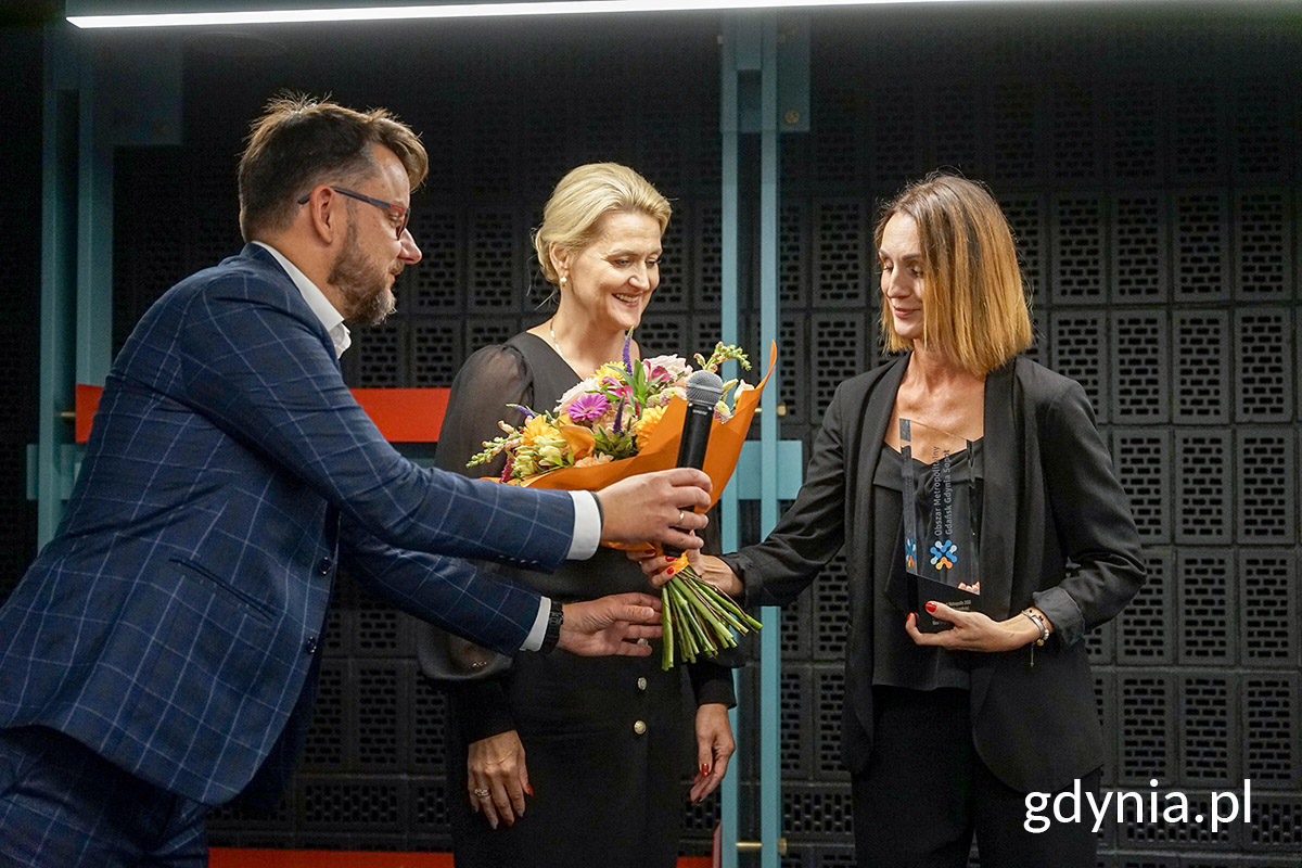 Sala konfernecyjna, trzy osoby na scenie, wręczają nagrodę