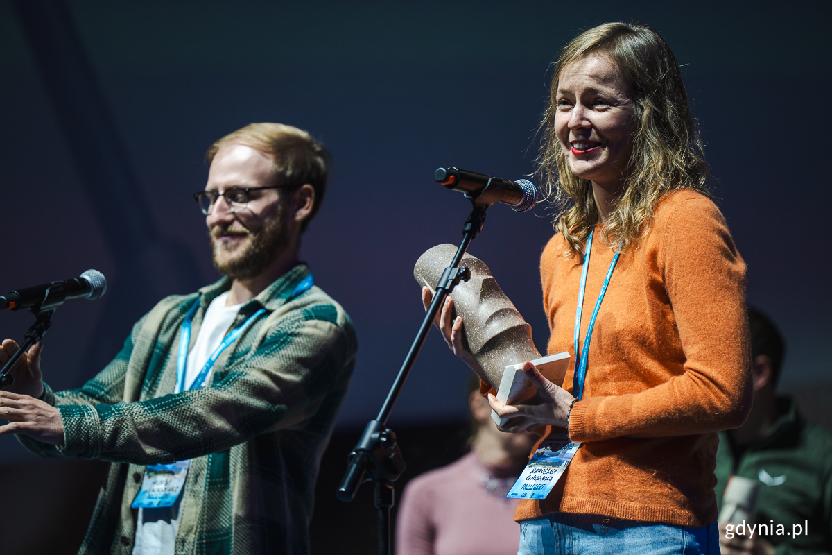 Młoda kobieta i młody mężczyzna na scenie z nagrodami podr&oacute;zniczymi.