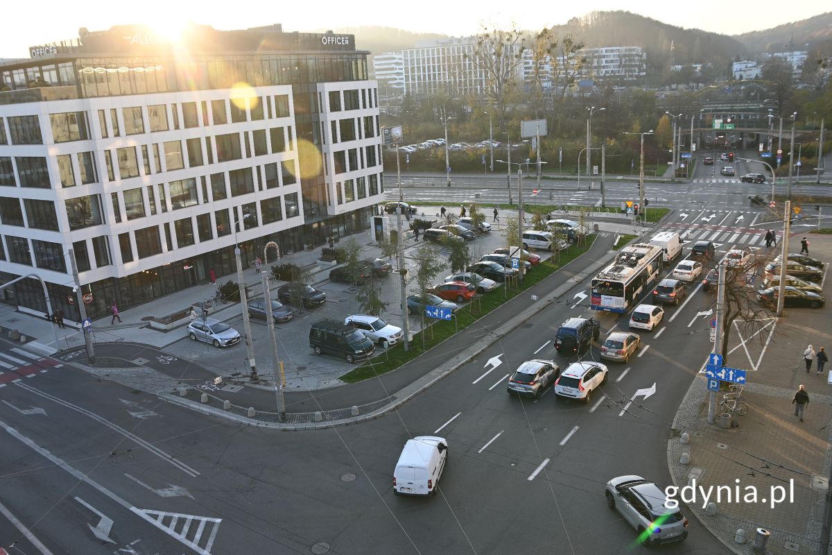 Dwa skrzyżowania i parking w widoku z g&oacute;ry, widoczne kolorowe samochody, piesi i ściany okolicznych budynk&oacute;w, a w oddali r&oacute;wnież drzewa