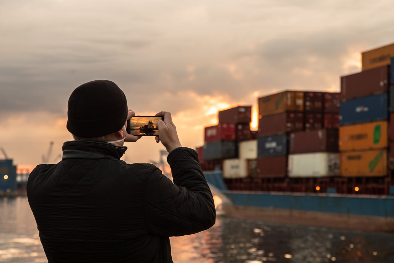 Statek z kontenerami wpływający do portu, zach&oacute;d słońca, mężczyzna w czapce robi zdjęcie smartfonem