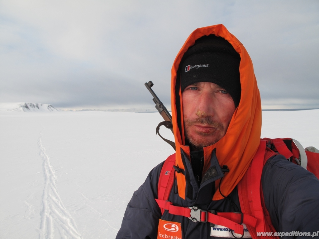 Mężczyzna w ziomowym ubraniu i z karabinem na plecach na śnieznym pustkowiu.