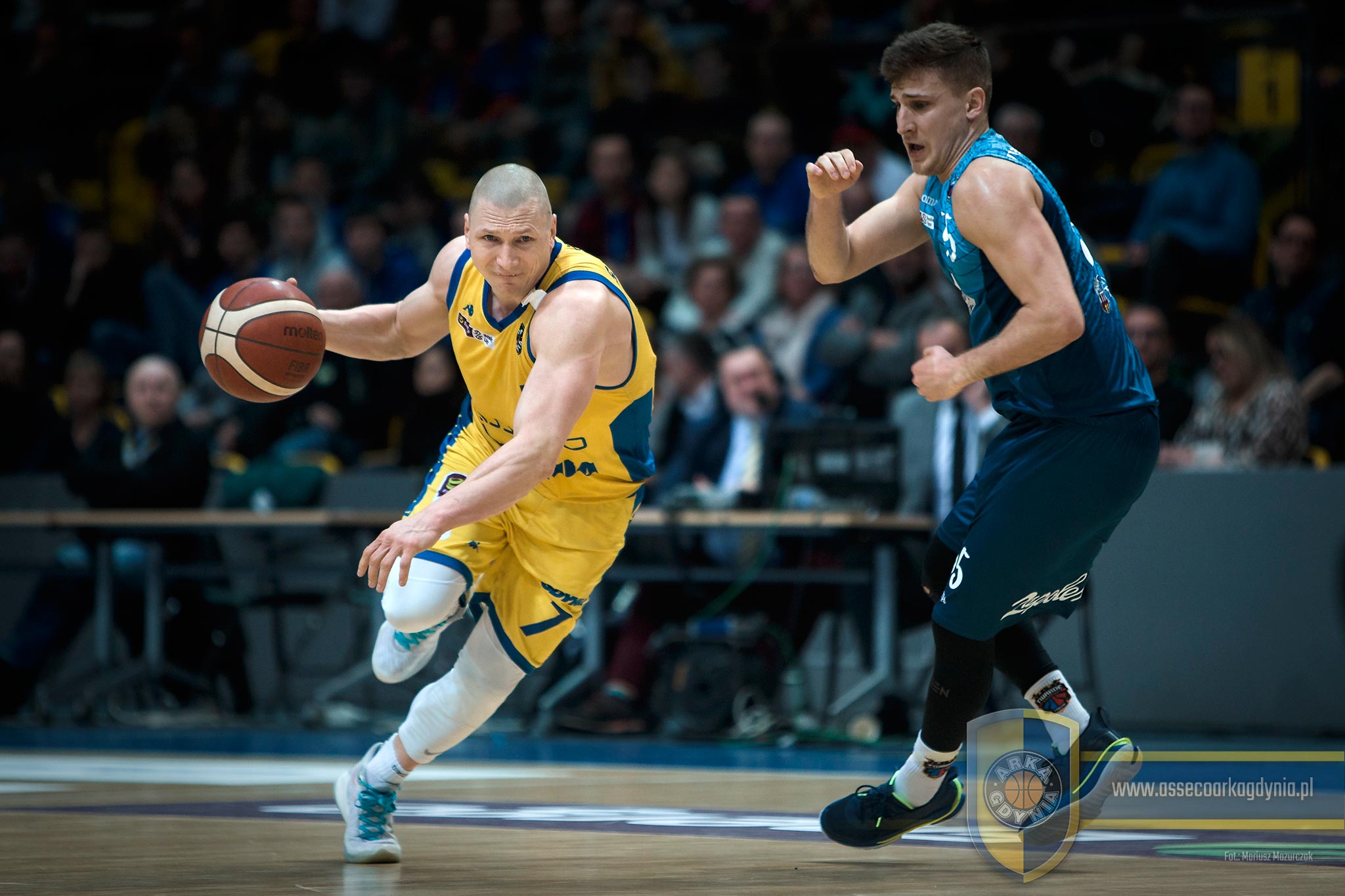 Na zdjęciu widzimy dw&oacute;ch koszykarzy, grających dla przeciwnych drużyn. W lewej części zdjęcia znajduje się zawodnik z piłką. Ma na sobie ż&oacute;łty str&oacute;j i kozłuję piłkę prawą ręką. Pr&oacute;buje ominąć koszykarza w granatowym stroju, kt&oacute;ry usiłuje zastawić drogę temu pierwszemu. Obaj znajdują się na drewnianym, lakierowanym parkiecie // fot. Mariusz Mazurczak (assecoarkagdynia.pl