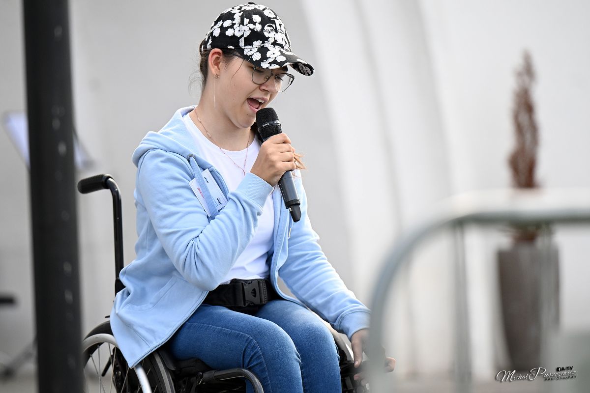 Dziewczyna ubrana w liliową bluzę, czapeczkę z daszkiem, siedząca na w&oacute;zku inwalidzkim , trzyma w ręku mikrofon