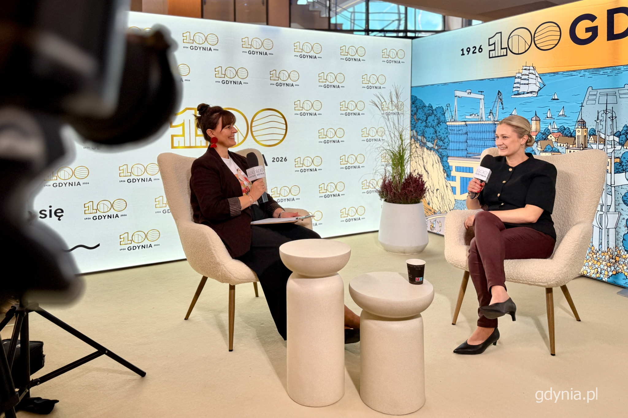 Studio "Gdynia Filmowa", Magdalena Śliżewska i Agata Kozierańska-Burda rozmawiają, siedząc w fotelach, w tle identyfikacja graficzna 100-lecia Gdyni