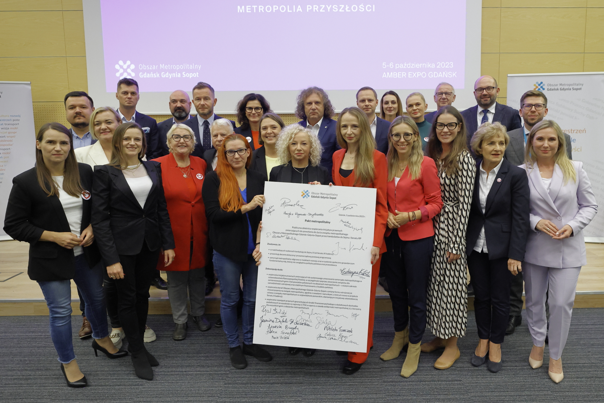 Grupa os&oacute;b pozuje do zdjęcia w sali konferencyjnej, trzymają dużą planszę z treścią Paktu Metropolitarnego