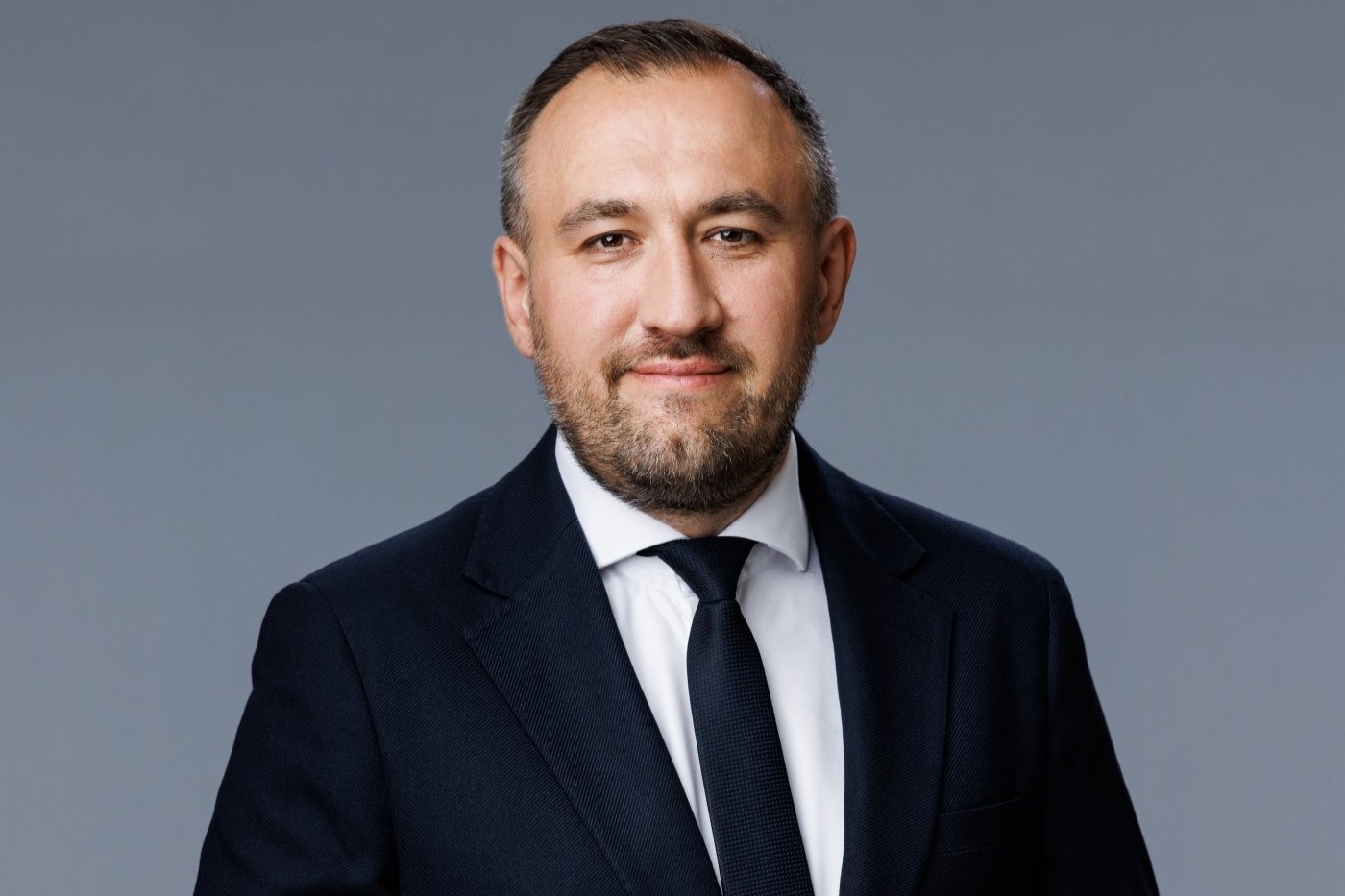 Tomasz Augustyniak pozuje do zdjęcia na białym tle, ubrany w ciemny garnitur i białą koszulę