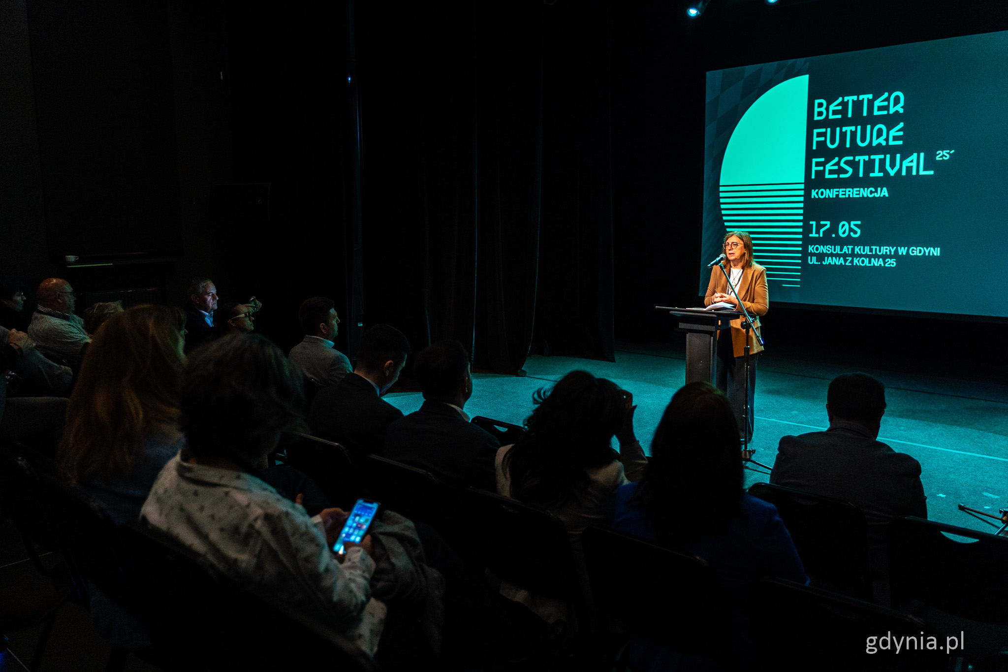 Sala w Konsulacie Kultury, przygaszone światło, ludzie na widowni, na scenie zielone podświetlenie, przemawia kobieta