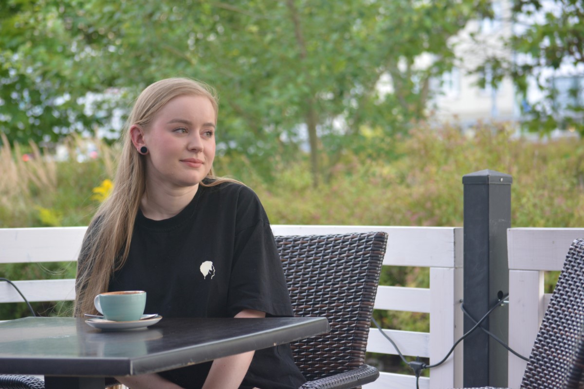 Zdjęcie przedstawia młodą kobietę siedzącą w restauracyjnym ogr&oacute;dku przy filiżance kawy. Wzrok kobiety skierowany jest na dalszy plan. 