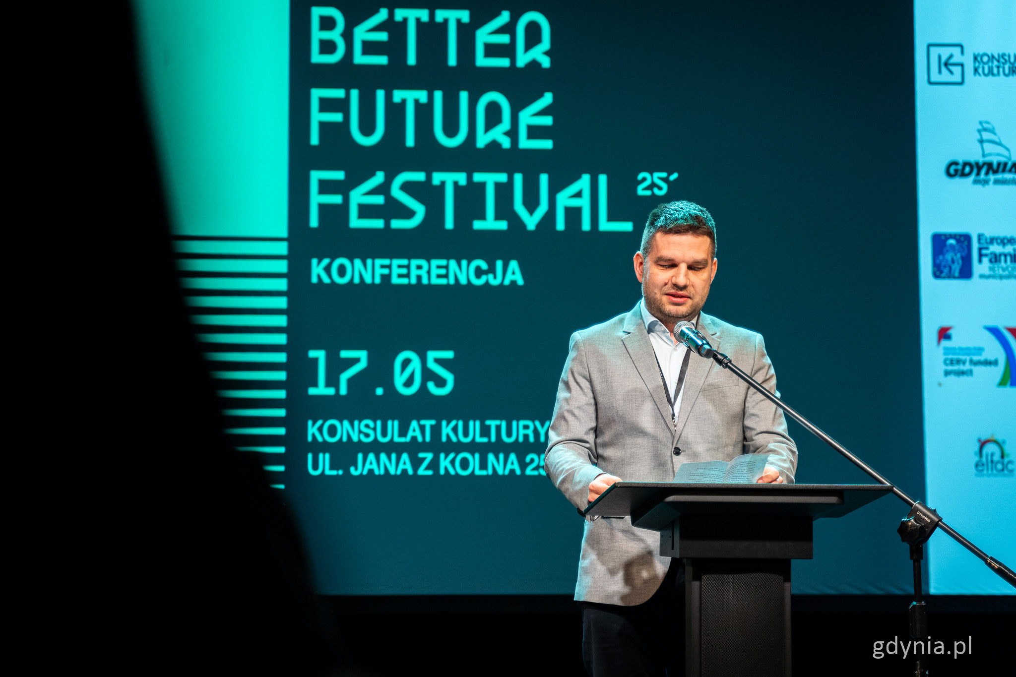 Sala w Konsulacie Kultury, Maciej Zielonka przemawia na scenie, za nim ekran z zielonym logotypem Better Future Festival