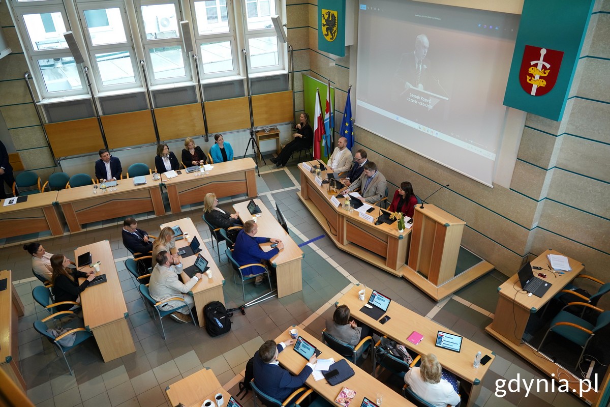 Sala obrad Rady Miasta Gdyni, radni siedzą w ławach, z boku tłumacz języka migowego, ekran z wyświetlaczem