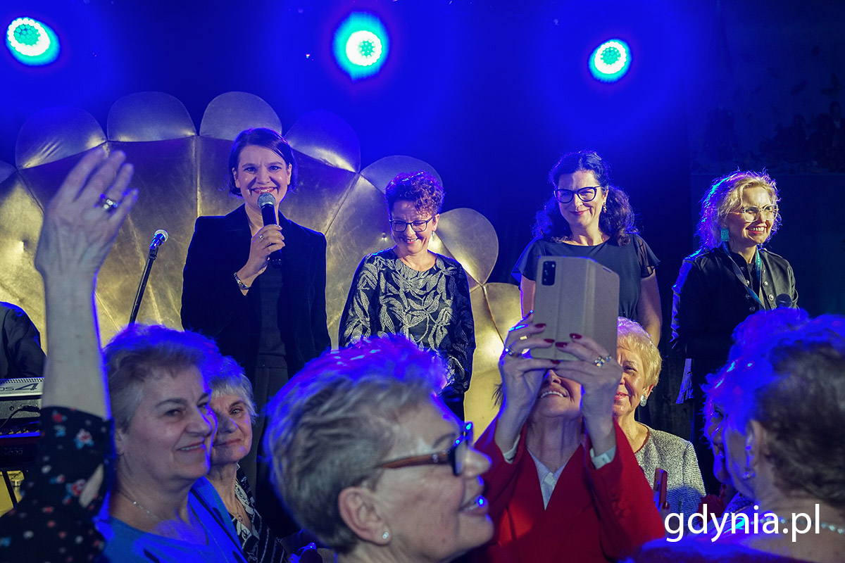 Seniorzy bawią się na balu na lotnisku, na scenie przemawiają do nich trzy kobiety.