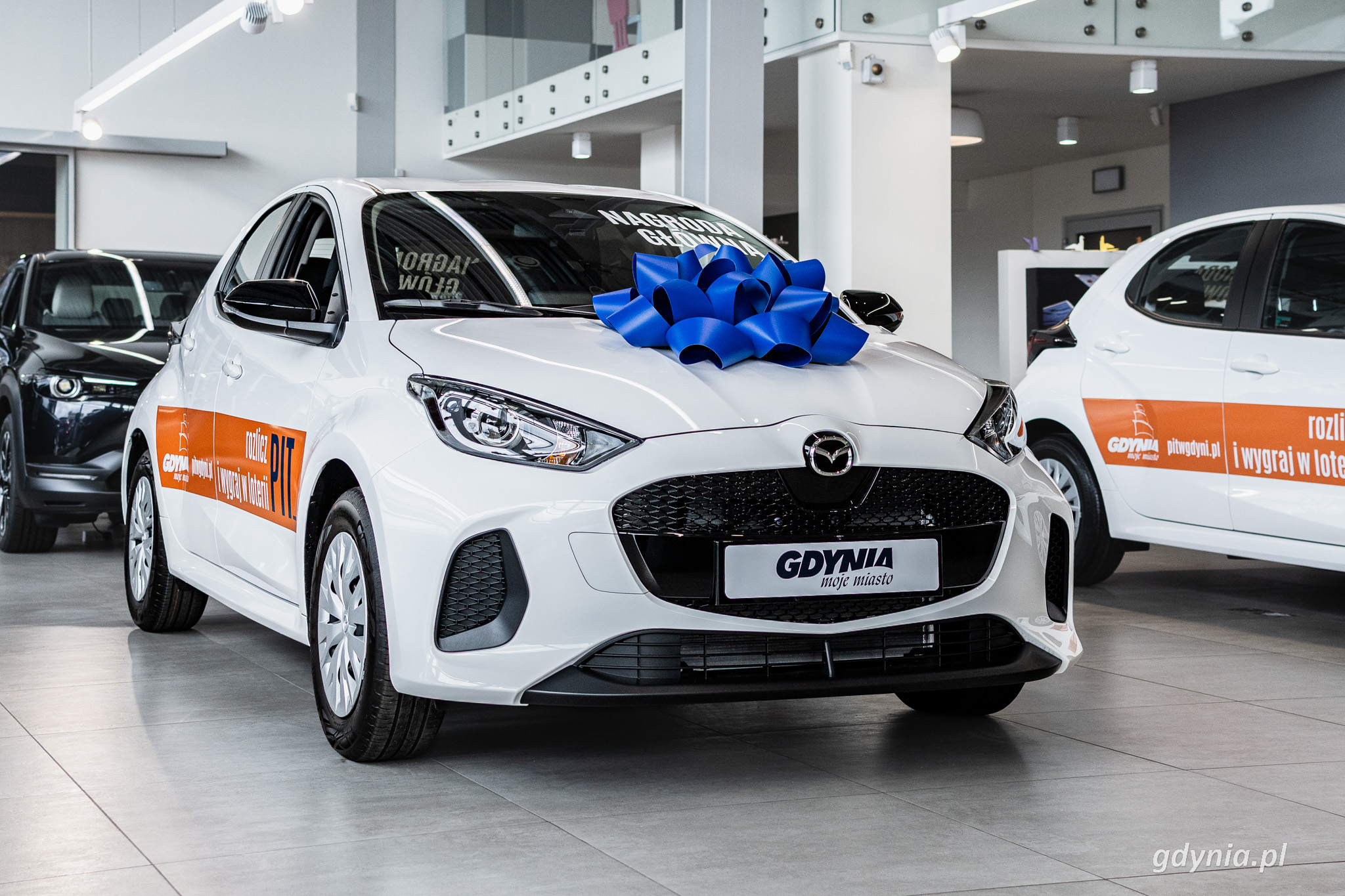 Biała Mazda 2, niebieska kokarda na masce, napis Nagroda Gł&oacute;wna na szybie, obok inne pojazdy, wnętrze salonu samochodowego