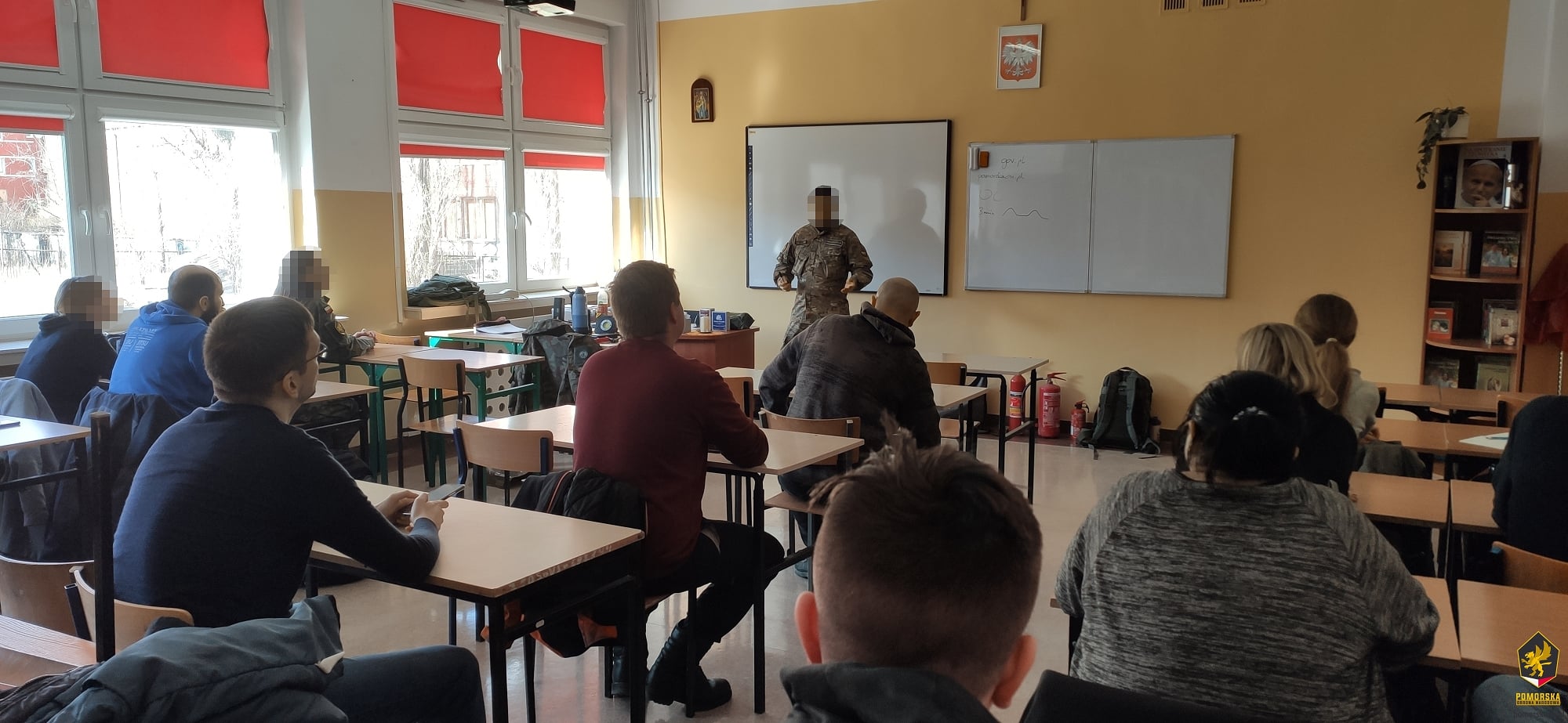 Szkolna sala, mężczyzna w mundurze na środku sali prowadzi zajęcia, w ławkach siedzą dorosłe osoby, kobiety i mężczyźni