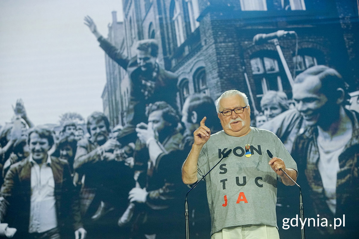 Lecha Wałęsa przemawia na scenie, w tle archiwalne zdjęcie ze strajk&oacute;w robotniczych