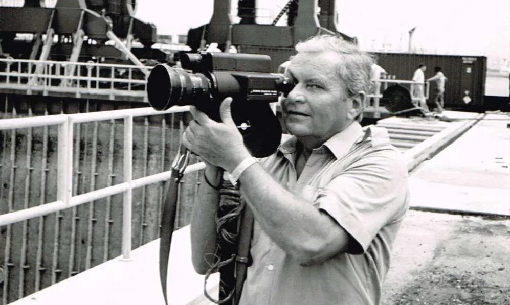 Czarno-białe archiwalne zdjęcie, Bohdan Sienkiewicz stoi przy nabrzeżu, trzyma przy oku kamerę i nagrywa, patrzy w obiektyw