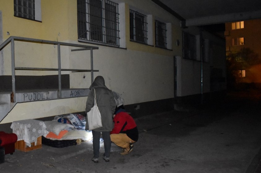 2.	Zdjęcie przedstawia dwoje pracownik&oacute;w MOPS podczas patrolu w nocy pochylających się nad osobą bezdomną kt&oacute;ra śpi na materacu rozłożonym pod schodami, przy tylnym wejściu do lokalu użytkowego. 