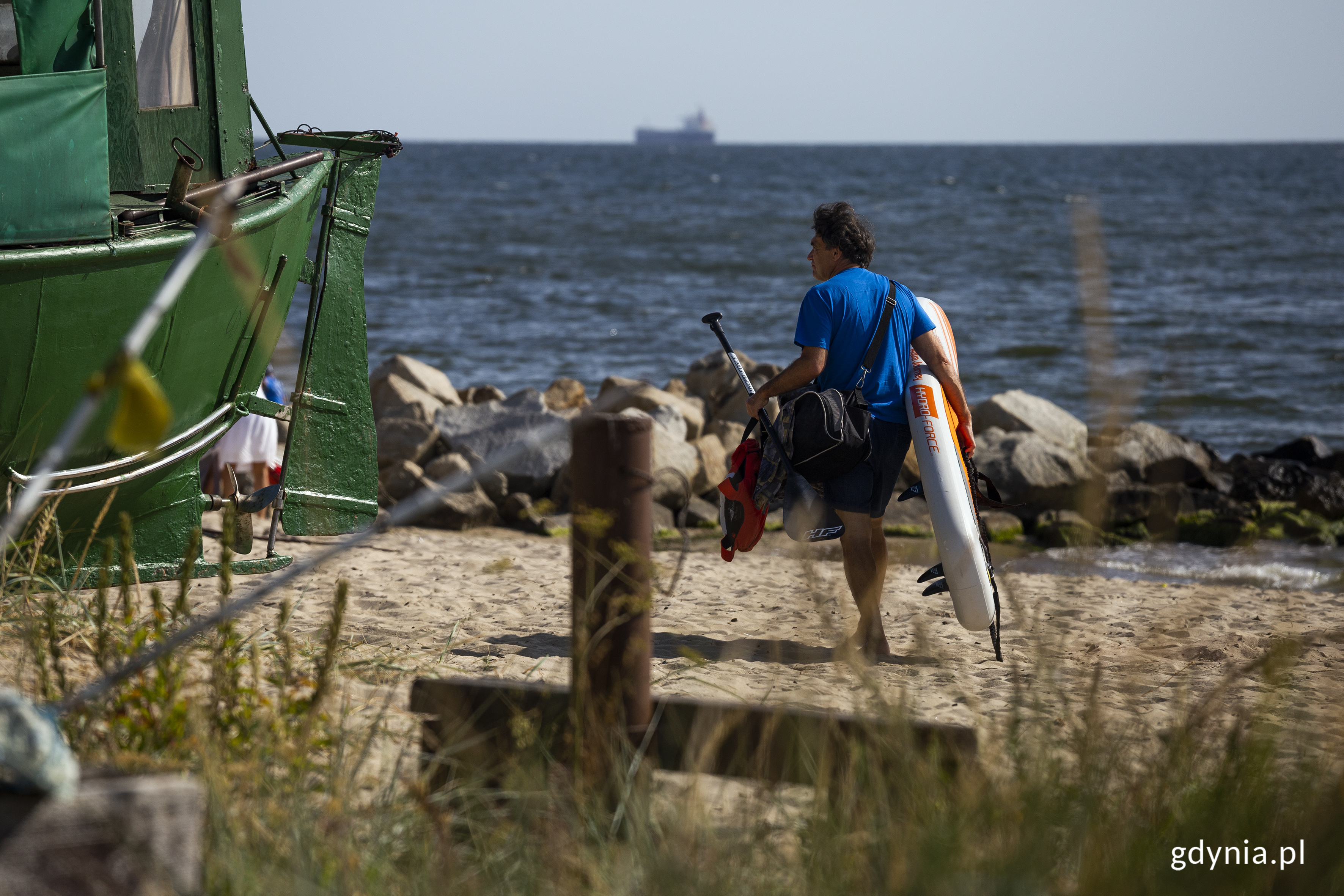 Plaża w Orłowie. Mężczyzna niesie deskę do pływania, obok kuter rybacki, w tle morze. Fot. Przemysław Kozłowski