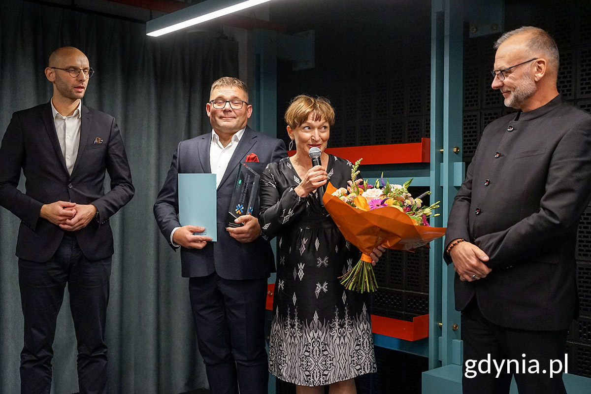 Sala konferencyjna, cztery osoby na scenie, dwie trzymają kwiaty i nagrody, kobieta przemawia przez mikrofon