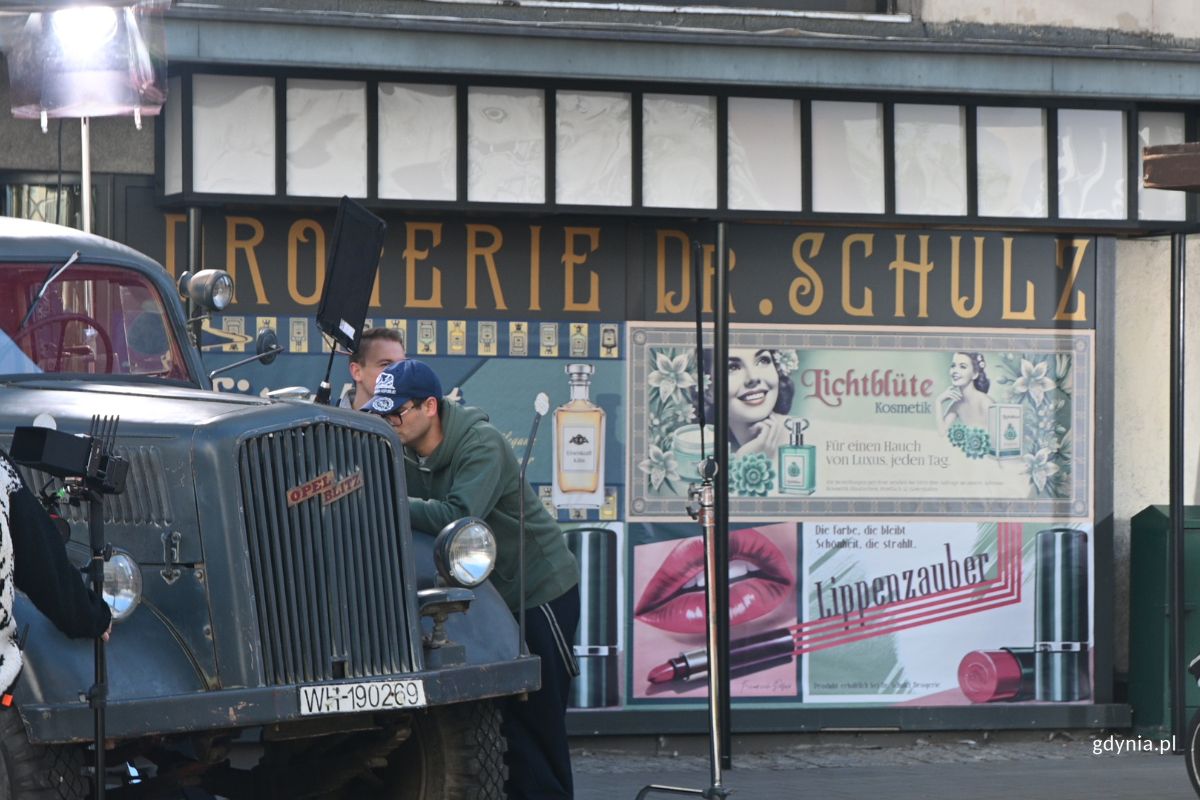 Niemieckojęzyczna witryna w oknie budynku i fragment szarej ciężar&oacute;wki, przy kt&oacute;rej stoją członkowie ekipy filmowej