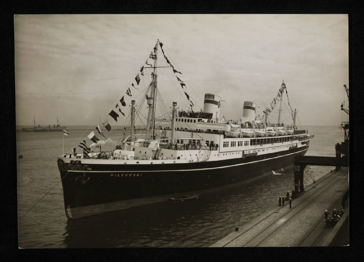 Czarno-białe zdjęcie statku pasażerskiego przy nabrzeżu z widoczną na dziobie nazwą &bdquo;Piłsudski&rdquo;