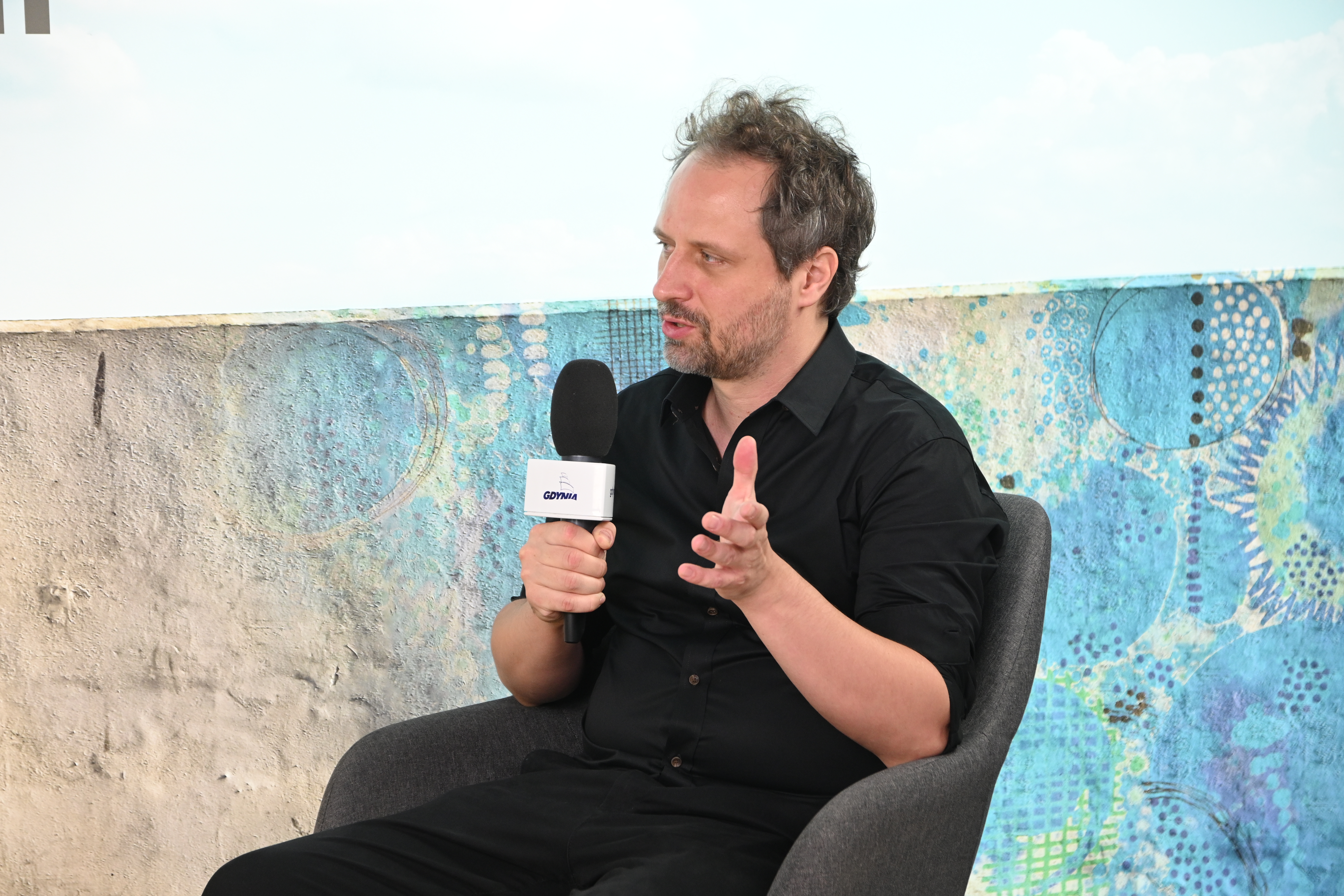 Studio telewizyjne, błękitne tło, siedzi Adrian Apanel, m&oacute;wi do mikrofonu