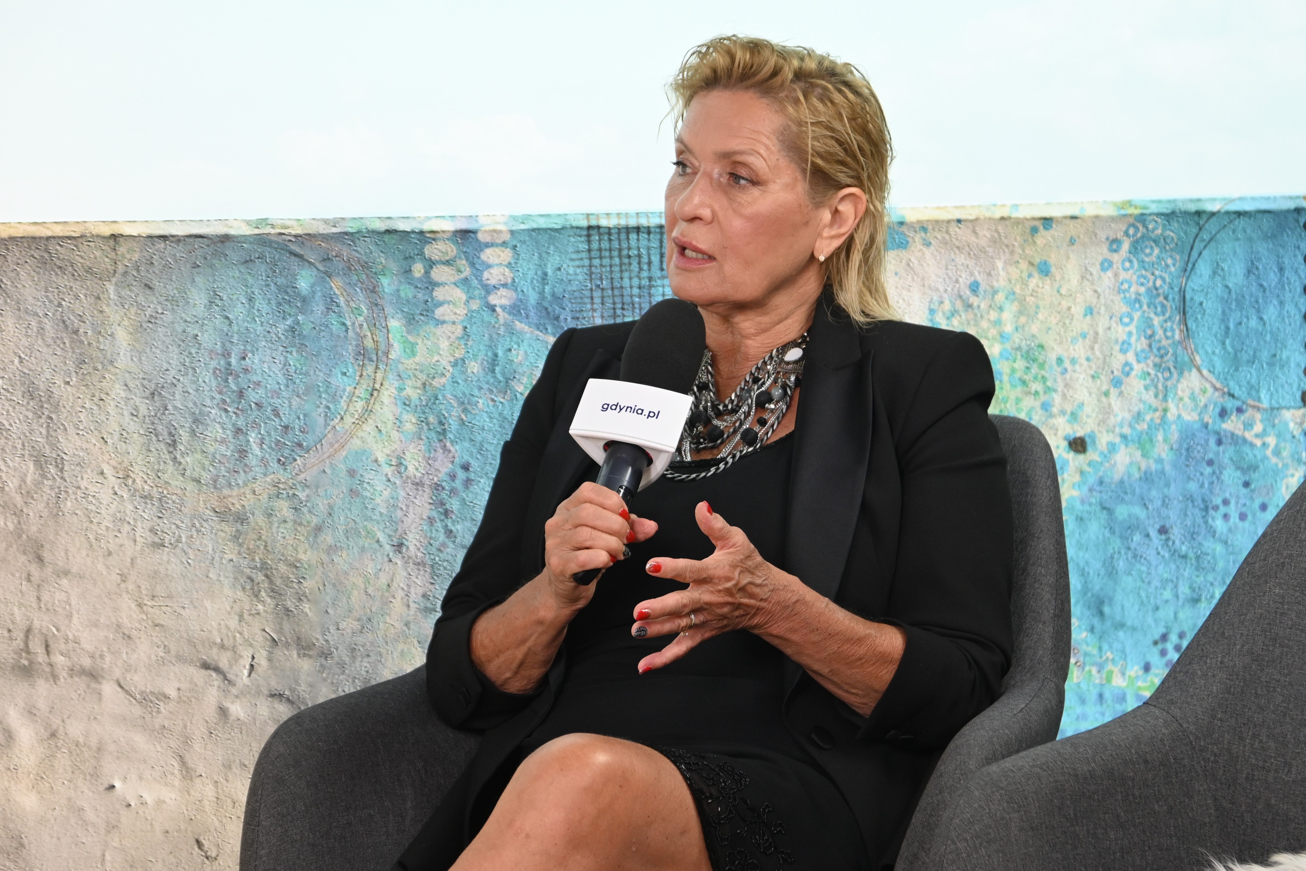 Ewa Kasprzyk siedzi w fotelu, m&oacute;wi do mikrofonu, za nią niebieskie tło