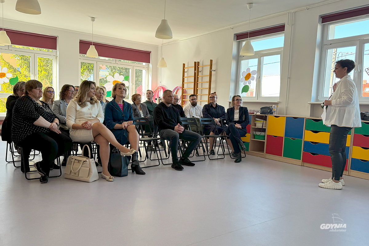Grupa os&oacute;b siedzących na krzesłach w sali przedszkolnej i słuchających stojącej przed nimi kobiety
