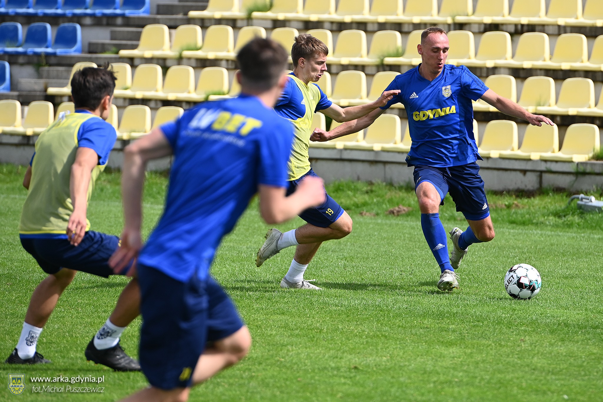 Na zdjęciu widzimy czterech biegnących piłkarzy w niebieskich strojach. Jeden z nich ma przy nodze piłkę i przymierza się do kopnięcia jej. Zawodnicy znajdują się na pokrytym zieloną murawą boisku. W tle widzimy puste ż&oacute;łto-niebieskie krzesełka trybun // fot. Michał Puszczewicz (Arka Gdynia SSA)