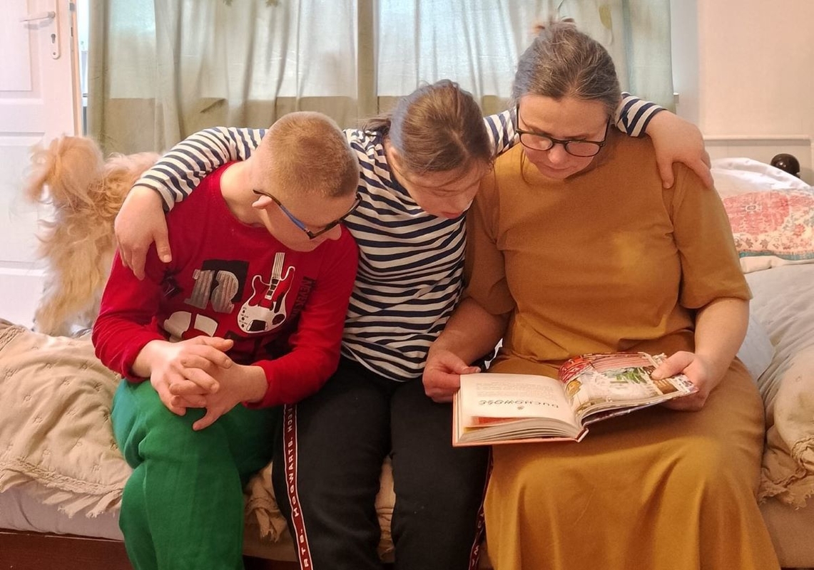 Zdjęcie przedstawia panią Beatę siedzącą na ł&oacute;żku i czytającą książkę dwojgu ze swoich dzieci