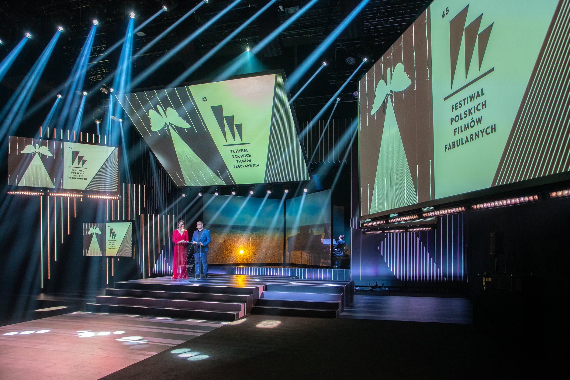 Zdjęcie z gali zamknięcia 45. Festiwalu Film&oacute;w Fabularnych. Oświetlona jasnymi reflektorami sala z kilkoma telebimami, na kt&oacute;rych widać logo festiwalu. Na dole, przy pulpicie - prowadzący Agata Konarska i Mateusz Cieślik // fot. Anna Rezulak