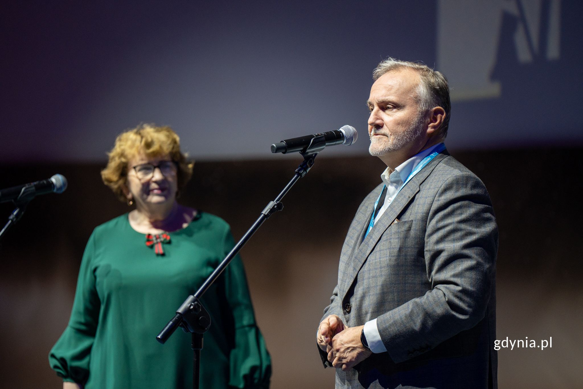 Mężczyzna w garniturze m&oacute;wi do mikrofonu. W tle kobieta.